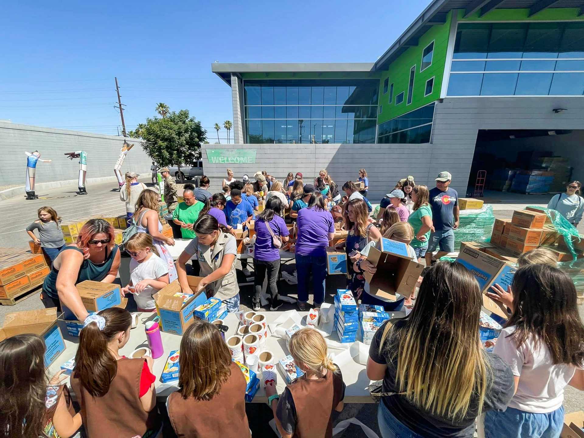large group of girl scouts and volunteers work together to pack boxes for care to share