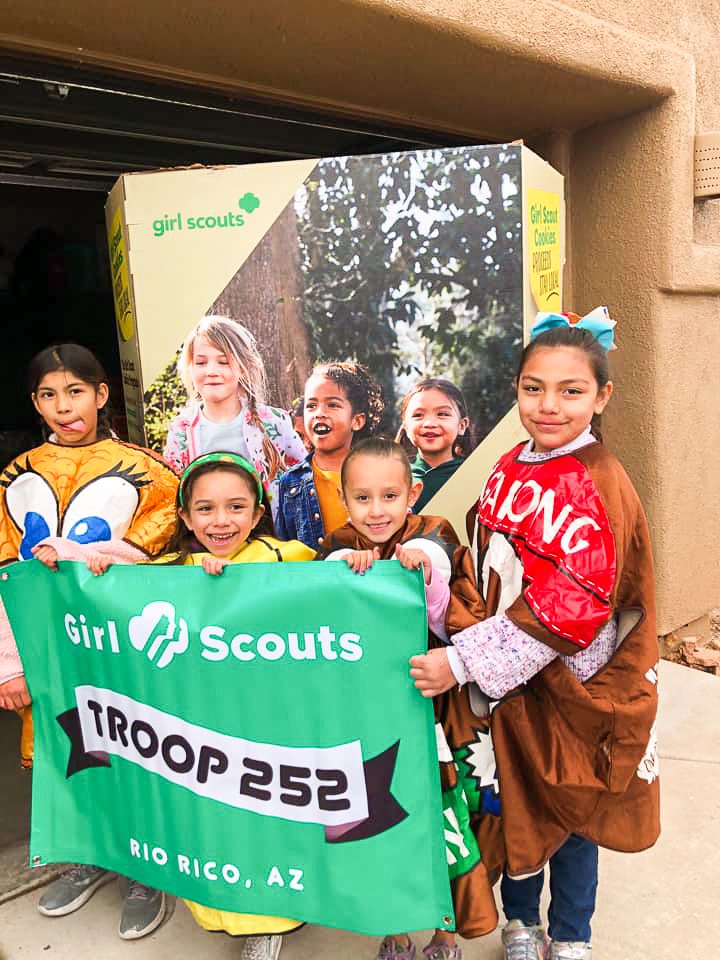 girl scouts holding up troop banner in front of large box of girl scout cookies