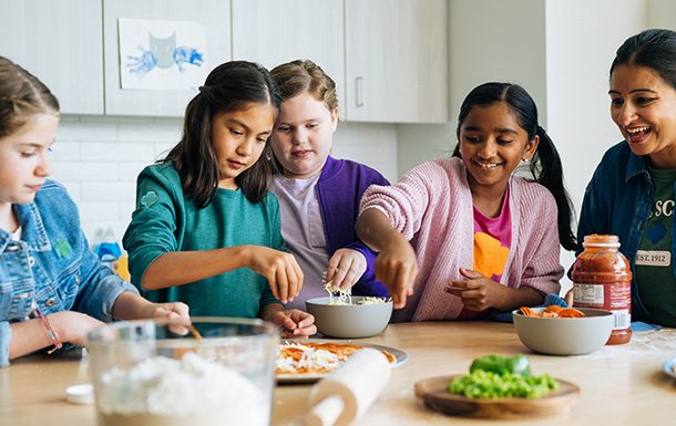 girl scouts with volunteers make a dish together